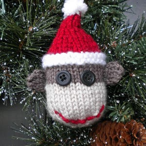 May include: A knitted sock monkey ornament wearing a red Santa hat with a white trim. The ornament is hanging on a green artificial Christmas tree with brown pine cones.