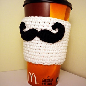 May include: White crocheted coffee cosy with a black felt moustache. The cosy is on a brown and orange takeaway coffee cup with the text "CAUTION HANDLE WITH CARE I'M HOT".
