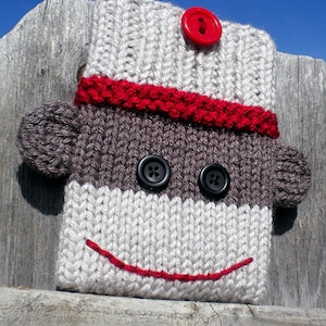 Może przedstawiać: Wełniana, dziana osłonka na urządzenie w kształcie małpy w skarpetce, z czerwonymi i białymi paskami, czarnymi guzikowymi oczami i czerwonym, haftowanym uśmiechem. Osłonka pasuje do Kindla, Nooka, iPada lub iPada Mini.