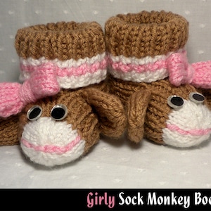 Può includere: Un paio di scarpine per neonati in maglia marrone con fiocchi rosa e dettagli bianchi. Le scarpine hanno la forma di scimmie con occhi a bottone neri.