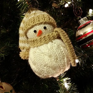May include: A knitted snowman ornament with a white body, brown hat and scarf, and black eyes. The ornament is hanging on a Christmas tree with white lights.
