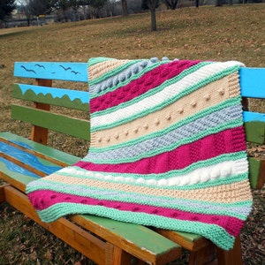 May include: A multi-colored striped baby blanket with different knit textures, including bobbles, is draped over a wooden bench with blue and green painted designs. "Treasured Sampler Baby Blanket" is written in the bottom of the image.