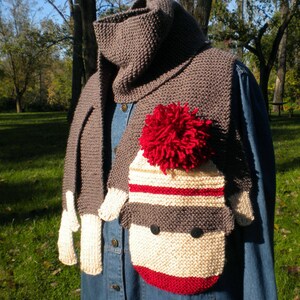 May include: A gray knitted scarf with a sock monkey design. The monkey has a red pom-pom hat, white and gray stripes, and red accents. The scarf is made of soft yarn and is perfect for keeping warm in the winter.