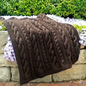 Könnte beinhalten: Eine braune gestrickte Kabelstrickdecke mit einem strukturierten Muster. Die Decke liegt auf einer Steinmauer mit grünen Pflanzen und lila Blumen.