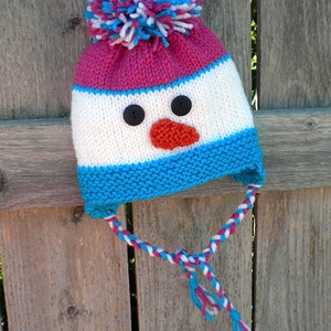 May include: A knitted snowman hat with a pink, white, and blue striped design. The hat has a white pom-pom on top and a black button nose and eyes. The hat has blue and white earflaps that tie together under the chin.