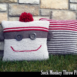 Op de afbeelding: Twee gebreide kussens, een met een sok-aap gezicht en een rode pompon, en de andere met grijze en rode strepen. De kussens liggen op een bed van groen gras voor een stenen muur. "Sock Monkey Throw Pillows" staat in zwarte tekst onderaan de afbeelding.