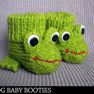 May include: A pair of green knitted baby booties shaped like frogs. The booties have black eyes with white pupils and red stitched smiles.