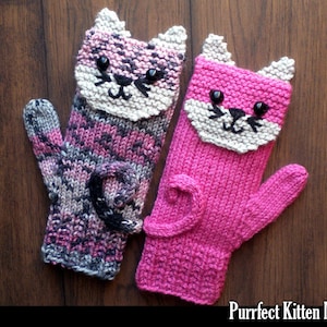 Könnte beinhalten: Ein Paar gestrickte Fäustlinge in Form von Katzen. Ein Fäustling ist rosa mit weißem Gesicht und schwarzen Details, der andere ist eine Mischung aus rosa, grau und schwarz mit weißem Gesicht und schwarzen Details. Beide haben einen eingerollten Schwanz.