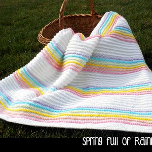 May include: A white crocheted baby blanket with colorful stripes in shades of pink, yellow, blue, and green. The blanket is folded and lying on a bed of green grass. The text "Spring full of Rainbows" is printed below the blanket.