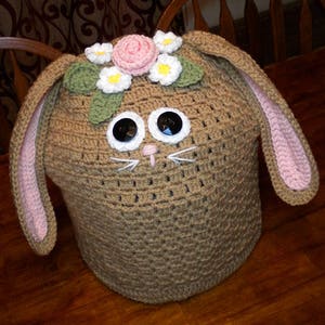 以下が含まれることがあります： ピンクの耳、茶色の毛皮、花冠が付いた、ウサギの形をした、かぎ針編みのインスタントポットカバー。花冠にはピンク、白、緑の花が飾られています。