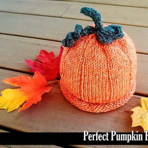 May include: A knitted orange pumpkin hat with a green stem and black leaves. The hat is sitting on a wooden surface with fall leaves.