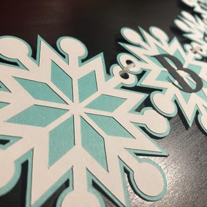 以下が含まれることがあります： 白、水色、黒を基調とした雪の結晶バナー。各雪の結晶は水色の中心部と白い縁取りが施されています。雪の結晶の一つには文字「B」が描かれています。バナーは紐で繋がれています。