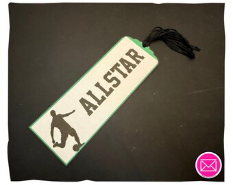 Puede incluir: Un marcapáginas rectangular con fondo color crema y borde verde. Presenta el texto negro ALLSTAR y una silueta negra de un jugador de fútbol pateando un balón. Una borla de hilo negro está unida a la esquina superior derecha.