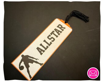 Puede incluir: Un marcapáginas rectangular con fondo color crema y borde naranja. La palabra ALLSTAR está impresa en letras mayúsculas negras sobre una silueta negra de un jugador de baloncesto botando un balón. Tiene una borla de hilo negro unida.