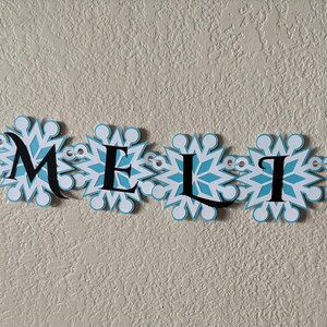 以下が含まれることがあります： 白と水色の雪の結晶のデザインの中に、黒い文字で「AMELIA」と書かれたバナー。 バナーには小さな透明な装飾が施されており、質感のある薄いベージュ色の壁に設置されています。