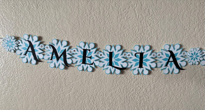 以下が含まれることがあります： 白と水色の雪の結晶のデザインの中に、黒い文字で「AMELIA」と書かれたバナー。 バナーには小さな透明な装飾が施されており、質感のある薄いベージュ色の壁に設置されています。