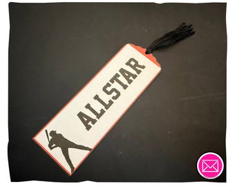 Puede incluir: Un marcapáginas rectangular con fondo color crema y borde rojo. La palabra ALLSTAR está impresa en letras mayúsculas negras grandes. Debajo del texto hay una silueta negra de un jugador de béisbol bateando. Tiene una borla de hilo negro unida.