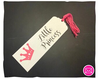 Puede incluir: Un marcapáginas rectangular de color blanco roto con el texto Little Princess en letras cursivas negras. Un gráfico de corona rosa se sitúa debajo del texto. El marcapáginas incluye un agujero en la parte superior con una borla de hilo rosa.