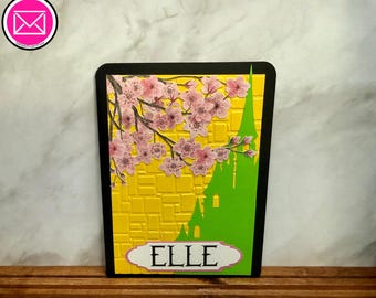 Puede incluir: Una tarjeta de felicitación rectangular con borde negro que presenta un fondo de ladrillo texturizado amarillo, una silueta verde de un castillo y ramas de cerezo en flor rosas. Una etiqueta blanca con borde rosa muestra el nombre ELLE en negro.