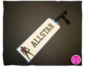 Puede incluir: Un marcapáginas rectangular con fondo beige claro y borde azul. La palabra ALLSTAR está impresa en letras mayúsculas negras en negrita. En la parte inferior hay una silueta negra de un jugador de fútbol americano con un balón. Un cordón negro está atado en la parte superior.