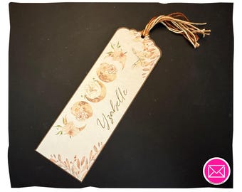 Puede incluir: Un marcapáginas rectangular con esquinas superiores redondeadas, fondo color crema y borde marrón. Muestra una serie de fases lunares decoradas con elementos florales, una ilustración de una polilla y el nombre Yzabelle en letra cursiva.