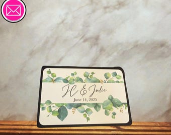 Puede incluir: Un letrero rectangular con borde negro y centro color crema. El diseño incluye ilustraciones de hojas de eucalipto verdes en la parte superior e inferior. El texto dice JC & Julie en letra cursiva, seguido de la fecha 14 de junio de 2025.