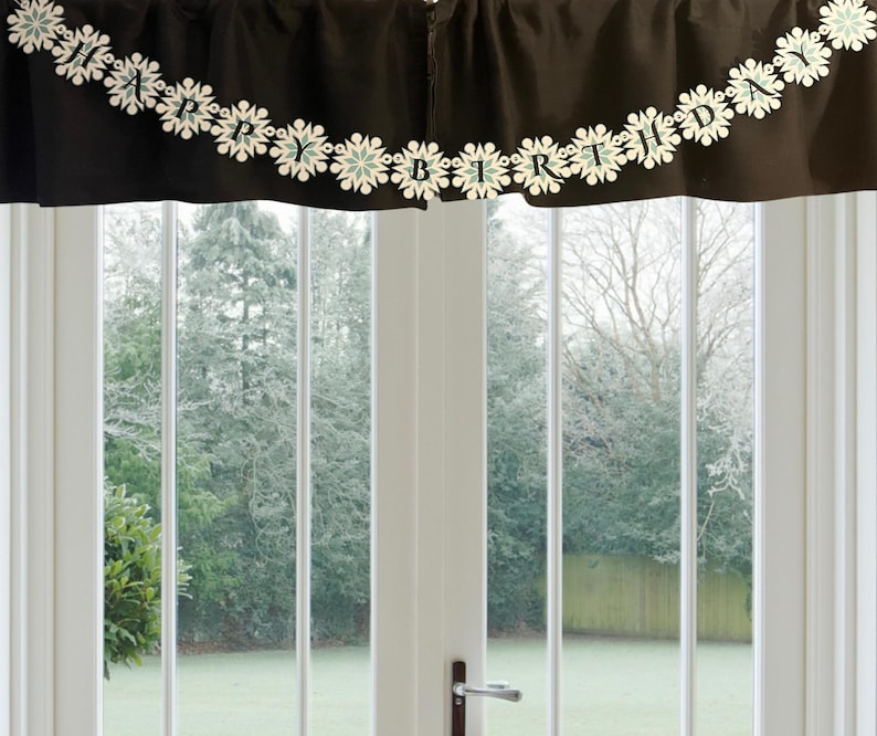 以下が含まれることがあります： 「HAPPY BIRTHDAY」と書かれた装飾的なガーランドが付いた黒い生地のバランス。ガーランドには白と水色の雪の結晶の切り抜きがあります。バランスは、緑の庭と木々が見えるガラスドアの上に吊り下げられています。