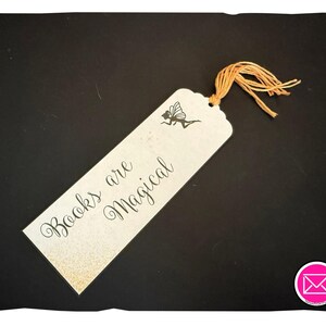 May include: A rectangular bookmark with a light cream background, featuring a black silhouette of a fairy and the text "Books are Magical" in black script. The bookmark has a scalloped top edge and a light orange tassel attached.