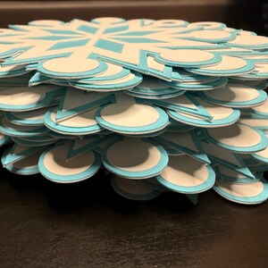 以下が含まれることがあります： 雪の結晶の形をした紙の切り抜きの山。各切り抜きは白で、水色のアクセントと水色の縁取りがあります。切り抜きは重ねられており、立体的な効果を生み出しています。雪の結晶のデザインは最上層に見えます。