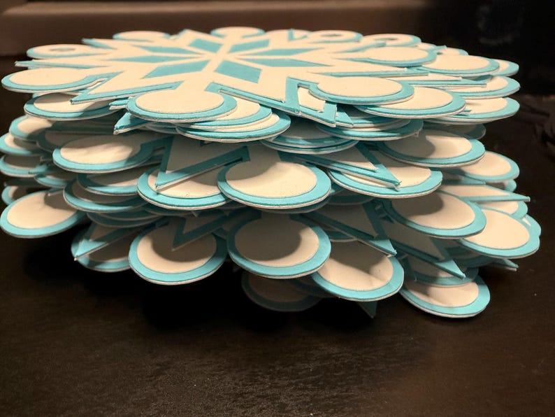 以下が含まれることがあります： 雪の結晶の形をした紙の切り抜きの山。各切り抜きは白で、水色のアクセントと水色の縁取りがあります。切り抜きは重ねられており、立体的な効果を生み出しています。雪の結晶のデザインは最上層に見えます。