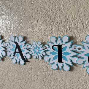 以下が含まれることがあります： 「AISLES」という単語が書かれたバナーが表示されています。各文字は黒い大文字で、白と水色の雪の結晶のデザインになっています。バナーは小さなアイレットで繋がれています。