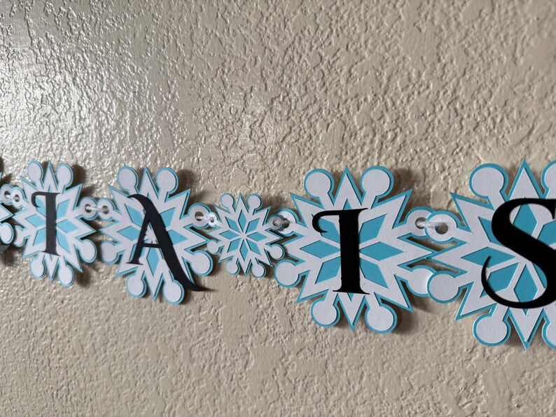 以下が含まれることがあります： 「AISLES」という単語が書かれたバナーが表示されています。各文字は黒い大文字で、白と水色の雪の結晶のデザインになっています。バナーは小さなアイレットで繋がれています。