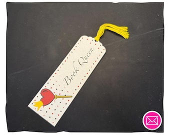 Puede incluir: Un marcapáginas rectangular de color blanco roto con el texto Book Queen en letra cursiva negra. El diseño incluye un patrón de pequeños corazones rojos y negros, un icono de corazón rojo con una corona amarilla y una borla de hilo amarillo.