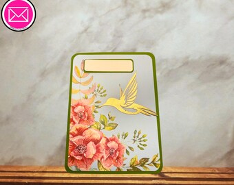 Puede incluir: Una tarjeta rectangular con borde verde que presenta la silueta de un colibrí dorado y flores rojas estilo acuarela con hojas verdes. La tarjeta incluye un espacio rectangular vacío y claro en la parte superior para texto o etiquetas.