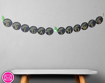Puede incluir: Una pancarta negra con letras doradas que dice "BRIDAL SHOWER". Cada letra está sobre un fondo circular negro con un diseño de hojas verdes. La pancarta está unida con hojas verdes.