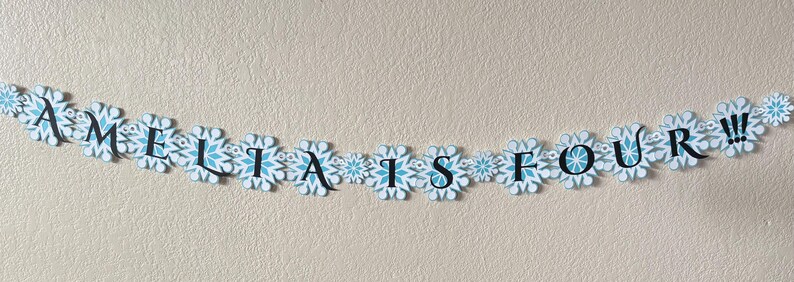 以下が含まれることがあります： 「AMELIA IS FOUR!!!」と書かれた、雪の結晶型の切り抜きで作られたバナー。雪の結晶は白と水色のアクセントと中心のデザイン。文字は黒色です。