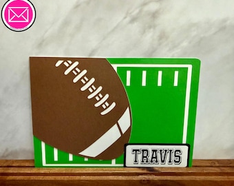 Puede incluir: Una tarjeta rectangular con un gráfico de balón de fútbol americano marrón con cordones y rayas blancas sobre un diseño de campo de juego verde brillante con líneas blancas. Una etiqueta blanca dice el nombre TRAVIS.