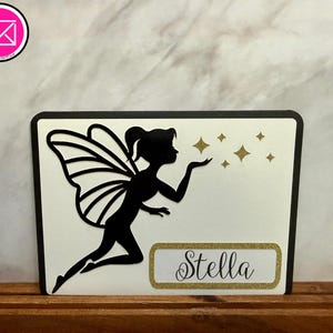 May include: A rectangular sign featuring a black silhouette of a fairy with wings blowing gold glitter stars. The sign has a white background with a black border and a gold glitter rectangular nameplate containing the name Stella in black script.