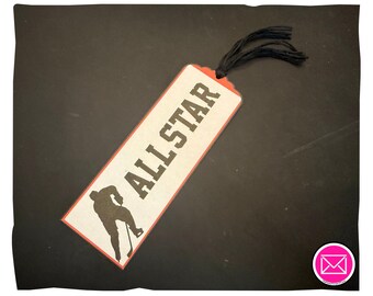 Puede incluir: Un marcapáginas rectangular con fondo color crema, que presenta el texto ALL STAR en letras negras en negrita y la silueta negra de un jugador de hockey. El marcapáginas tiene un borde naranja fino y una borla de hilo negro en la parte superior.