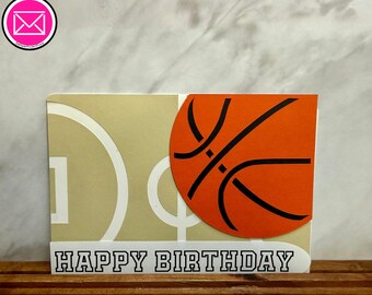 May include: A rectangular greeting card featuring a tan background with white basketball court line graphics. An orange basketball cutout with black lines is layered on the right side. The bottom white border displays the text HAPPY BIRTHDAY.