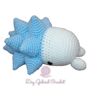 Puede incluir: Un juguete de peluche tejido a crochet con forma de criatura azul y blanca con pinchos. El juguete tiene un cuerpo blanco con una espalda azul con pinchos y un pequeño ojo negro.