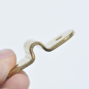 May include: A gold-colored metal bracket with a curved shape. The bracket has a small hole on the top.