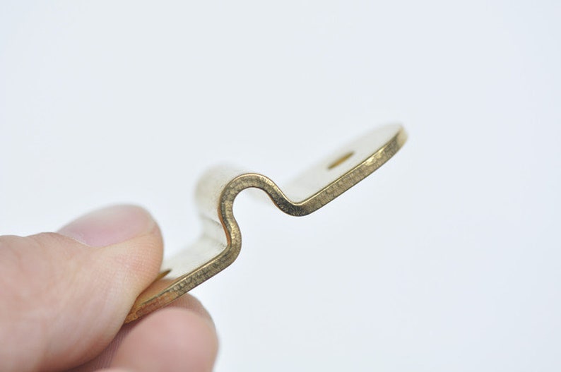May include: A gold-colored metal bracket with a curved shape. The bracket has a small hole on the top.