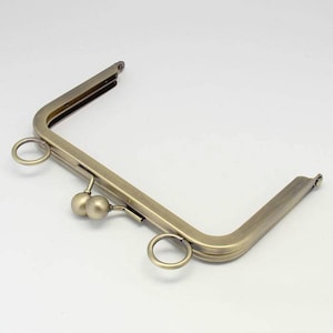 Op de afbeelding: Een gouden metalen tasframe met twee ronde ringen en een decoratieve bal sluiting.