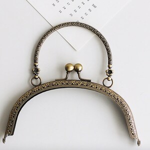 May include: A gold-toned metal purse frame with an ornate design and a top handle. The frame is designed to be used for a small purse or clutch.