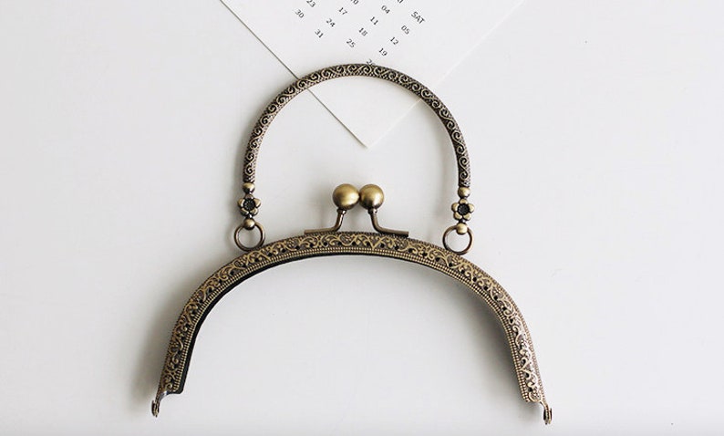 May include: A gold-toned metal purse frame with an ornate design and a top handle. The frame is designed to be used for a small purse or clutch.
