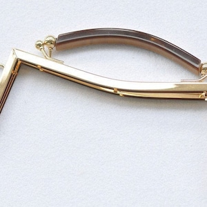 Puede incluir: Un marco de bolso de metal dorado con un asa curva marrón. El marco tiene forma rectangular con esquinas redondeadas y pequeños lazos para sujetar a una bolsa. El asa es de color carey.