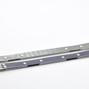 Könnte beinhalten: Zwei Metallarmbänder, eines silberfarben und eines dunkelblau, mit silbernen Nieten. Die Armbänder sind in der Mitte gefaltet und auf einer weißen Oberfläche ausgelegt.