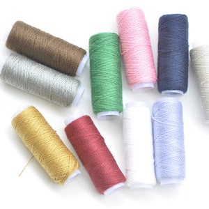 Può includere: Assortimento di bobine di filo colorate in varie tonalità di marrone, verde, rosa, blu, bianco, oro e rosso. Le bobine sono disposte in un cerchio su uno sfondo bianco.