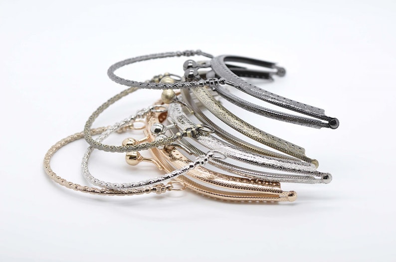 May include: A stack of six different metal purse frames. The frames are in various finishes, including silver, gold, and black. The frames are all in the shape of a half-circle and have a decorative design.